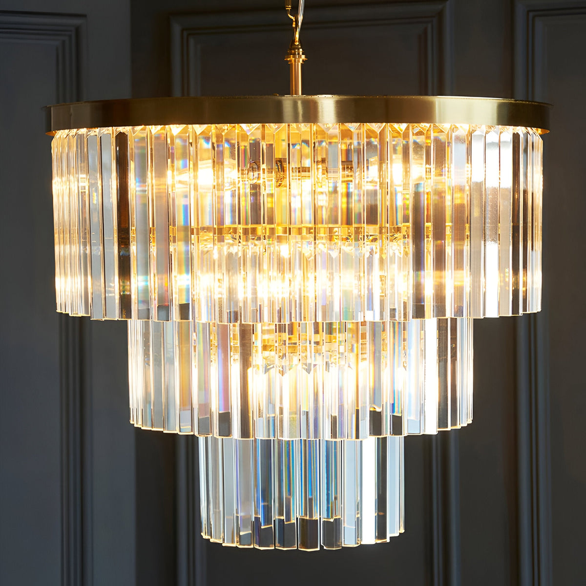 Endon Bevstead 7 light pendant in brushed brass with tiered clear cut lozenge glass drops, suspended from a chain against a grey panelled wall.
