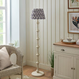 Endon Bevbury floor lamp with turned white spindle base and grey zigzag pleated shade, beside a wicker chair and cream sideboard in a bright living room.