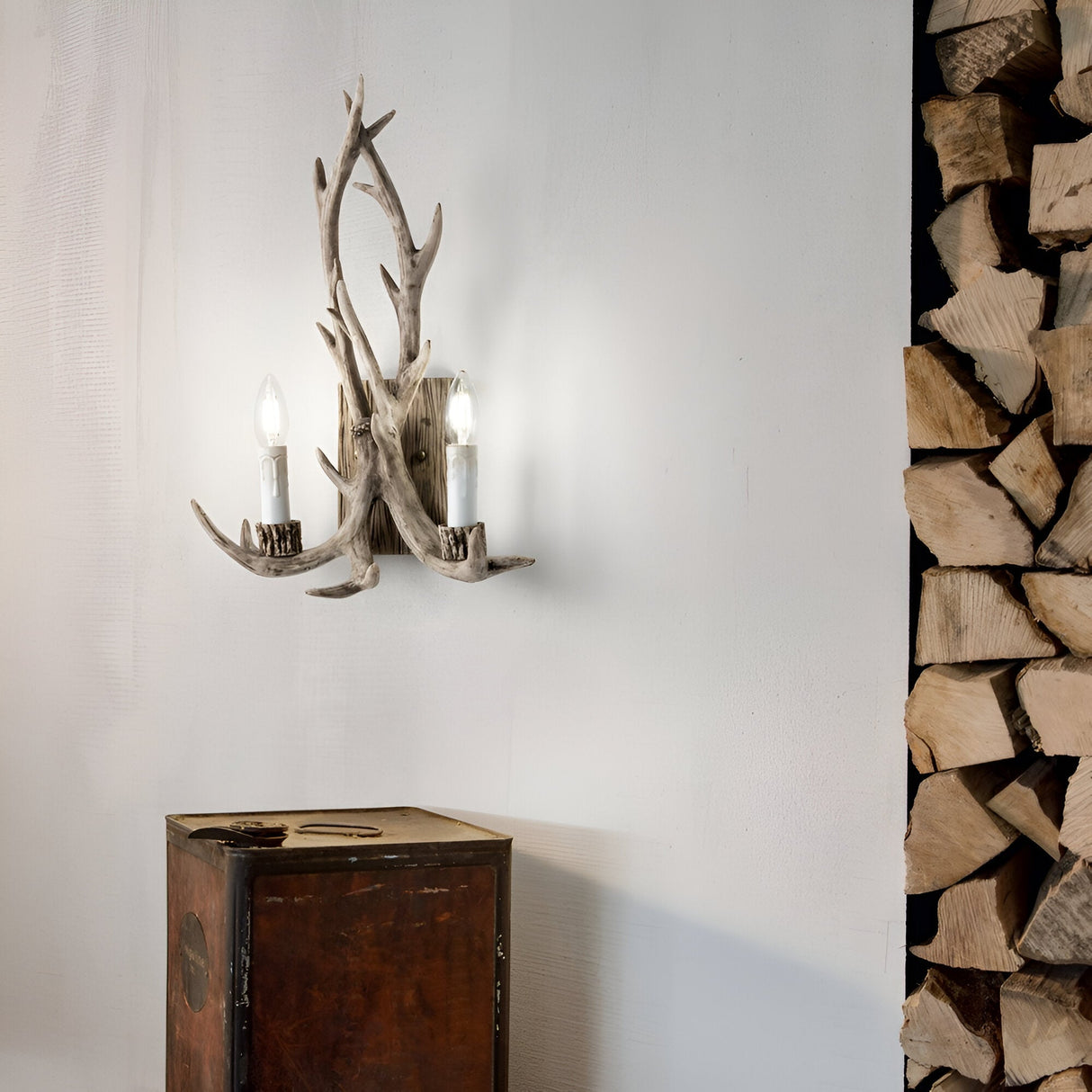 A Flow 2 Light Wall Light in Dark Brown with an antler-like design and two lit bulbs illuminates a white wall. Below it sits a wooden cabinet, enhancing the allure of this nature-inspired lighting. On the right, there's a neatly stacked pile of firewood.