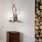 A Flow 2 Light Wall Light in Dark Brown with an antler-like design and two lit bulbs illuminates a white wall. Below it sits a wooden cabinet, enhancing the allure of this nature-inspired lighting. On the right, there's a neatly stacked pile of firewood.