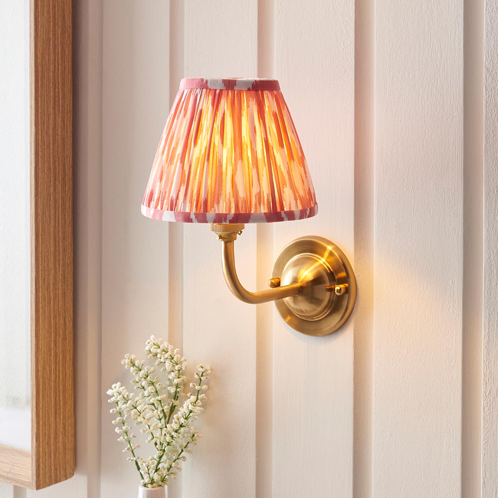 Endon Brackdale Ikat wall light with brushed aged brass curved arm and pleated coral pink Ikat shade, mounted on panelled wall above white flowers.