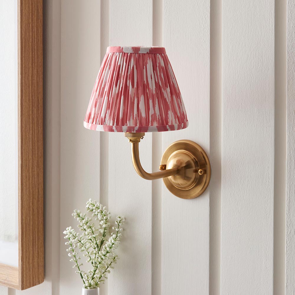 Endon Brackdale Ikat wall light with brushed brass curved arm and pleated coral pink Ikat shade, mounted on a white panelled wall above white blossoms.