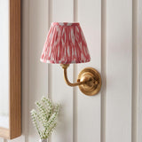 Endon Brackdale Ikat wall light with brushed brass curved arm and pleated coral pink Ikat shade, mounted on a white panelled wall above white blossoms.