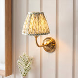 Brackdale Leaf wall light with brushed brass curved arm and pleated green leaf-print fabric shade, mounted on a panelled wall above white flowers.