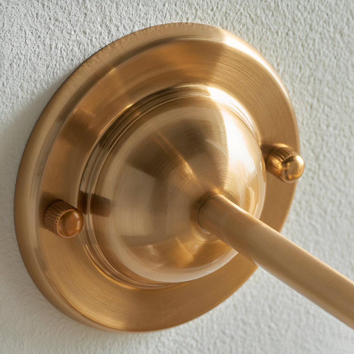 Close-up of the Brackdale Leaf wall light's dome-shaped brass base and curved arm, mounted on a textured white wall.