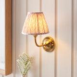Brackdale Leaf Wall Light in brushed brass with a curved arm and pleated fabric shade, mounted on a panelled wall beside a framed mirror and vase of flowers.