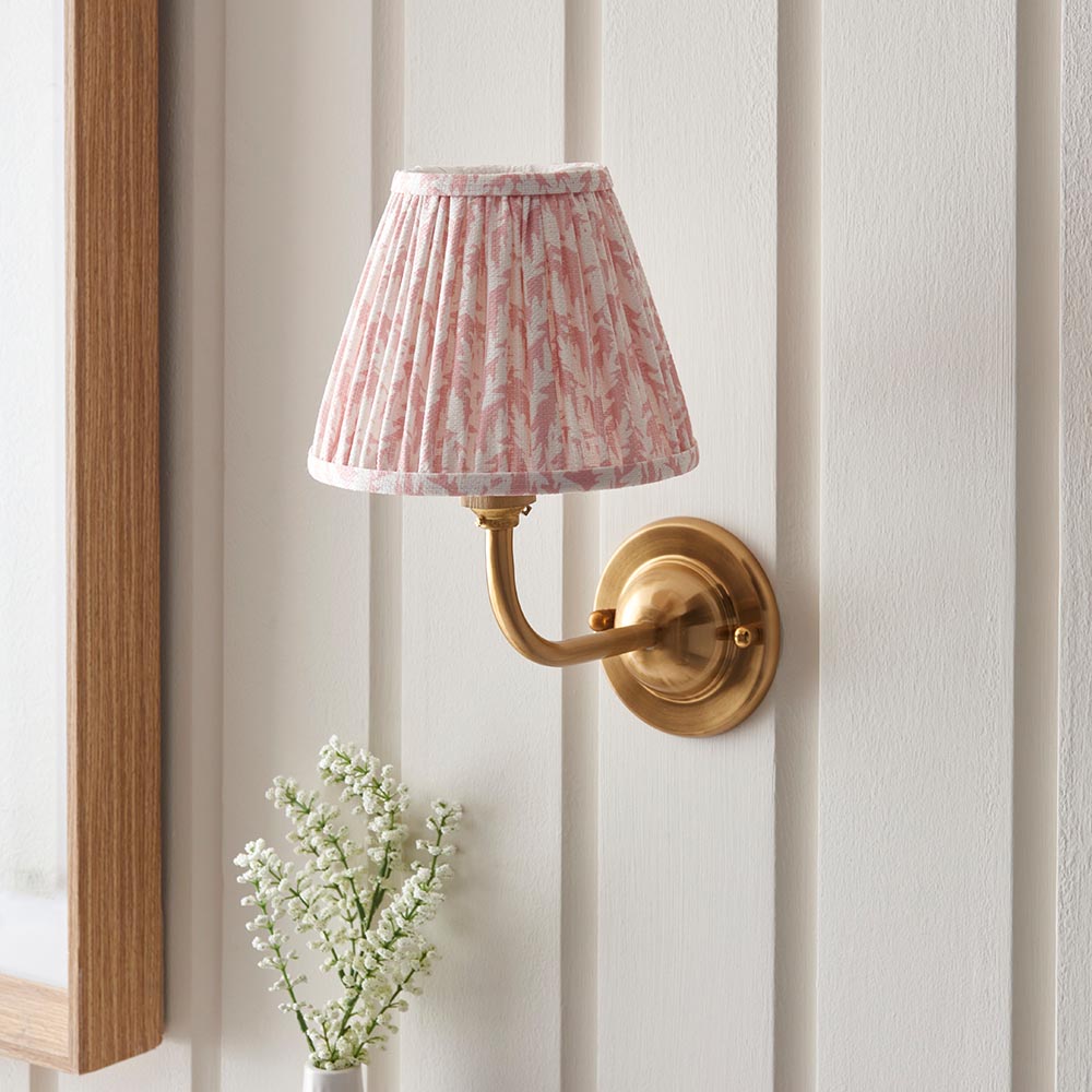 Brackdale Leaf Wall Light in brushed brass with a pink leaf-print pleated fabric shade, mounted on a panelled wall beside a mirror and flowers.