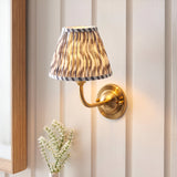 Endon Brackdale wall light in brushed aged brass with a pleated grey ripple faux linen shade, mounted on panelled wall beside a wooden mirror frame.