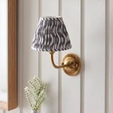Endon Brackdale wall light in brushed aged brass with a pleated grey ripple print tapered shade, mounted on white panelled wall beside a wooden mirror frame.