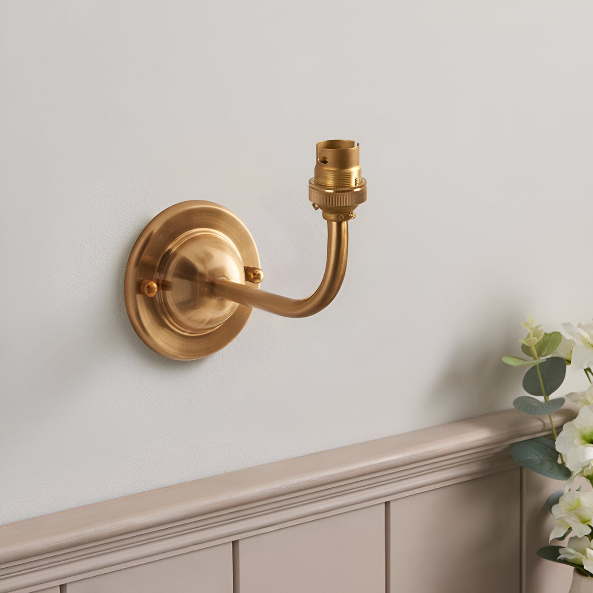 Endon Brackdale wall light base in brushed aged brass with curved arm and B22 lamp holder, mounted on a pale wall above grey panelling.