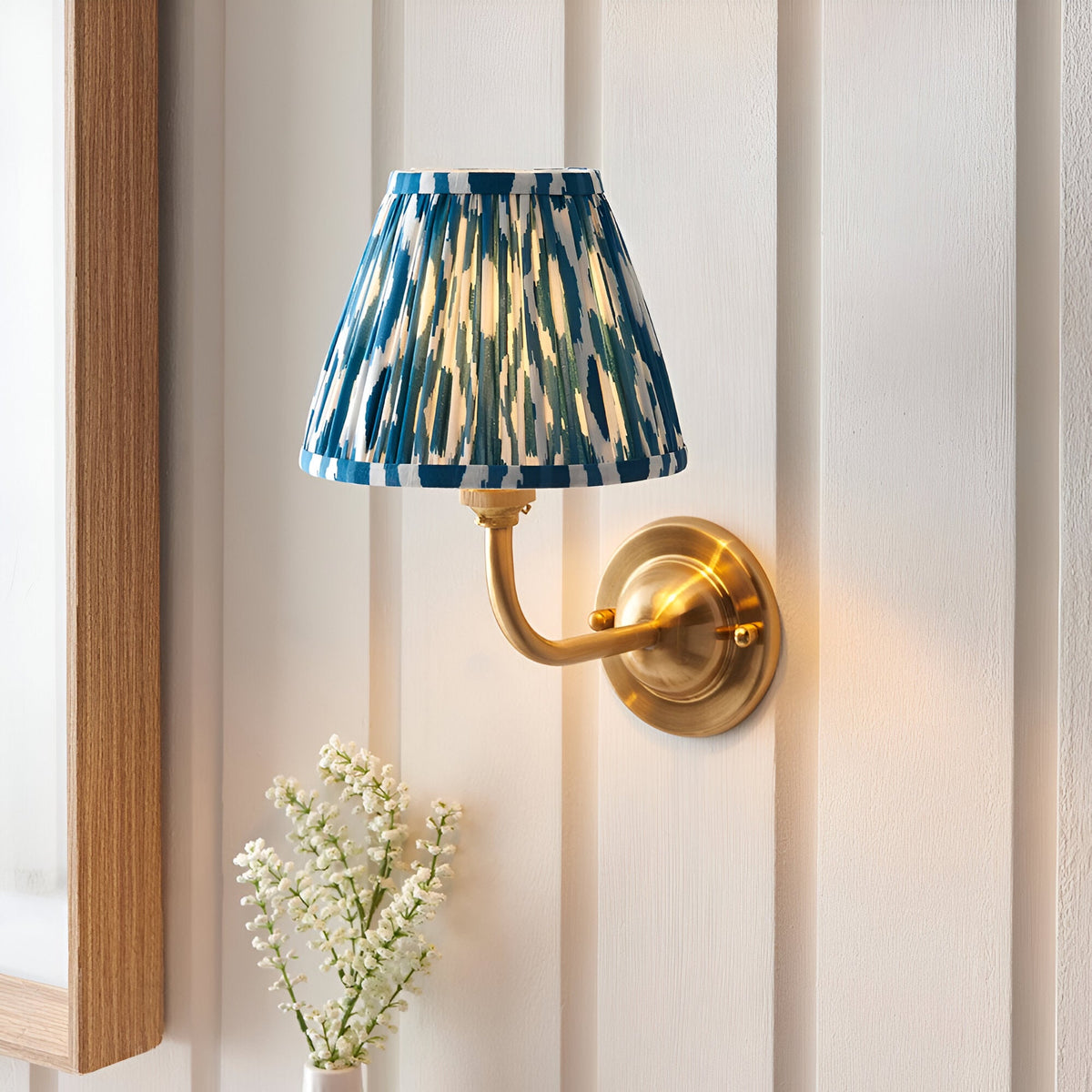 Endon Brackdale Ikat wall light in brushed brass with a pleated blue ikat fabric shade, mounted on white panelled wall above small white flowers.