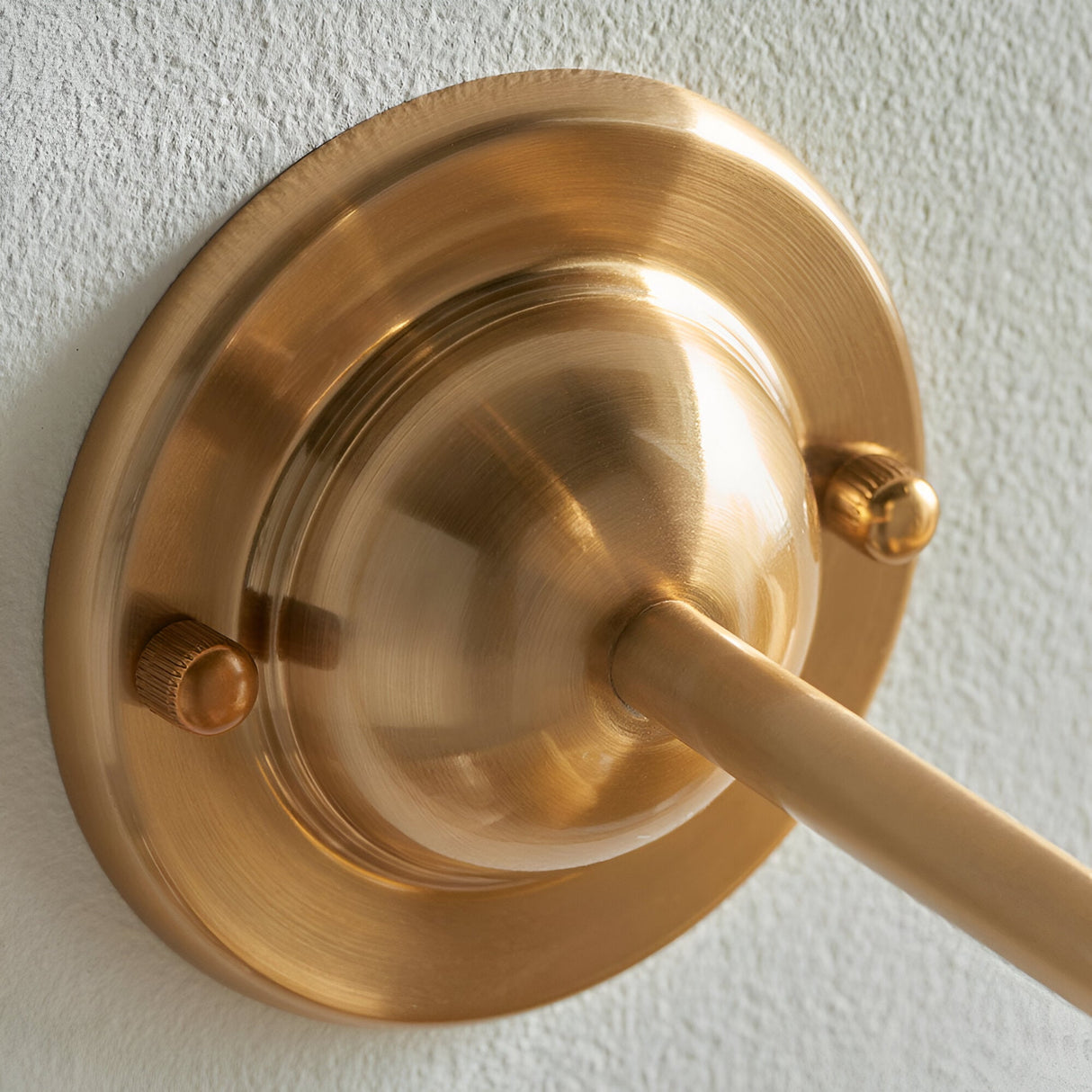Close-up of the Endon Brackdale Ikat wall light's brushed aged brass backplate and curved arm, mounted on a white textured wall.