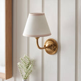 Brackdale Ivy wall light with brushed aged brass curved arm and tapered vintage white fabric shade, mounted on a white panelled wall above flowers.