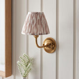 Endon Brackdale Ikat wall light with a brushed aged brass curved arm and dome, topped with a pleated pink and natural Ikat shade, mounted on panelled wall.