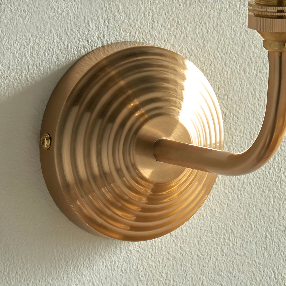 Close-up of the Endon Castgrove Ripple wall light's stepped, layered backplate and curved arm in brushed aged brass, mounted on a pale textured wall.