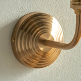 Close-up of the Endon Castgrove Ripple wall light's stepped, layered backplate and curved arm in brushed aged brass, mounted on a pale textured wall.
