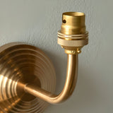 Close-up of Endon Castgrove Ripple wall light showing brushed aged brass curved arm, stepped backplate and B22 bayonet lamp holder against a white wall.
