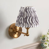 Endon Corven wall light with brushed brass adjustable arm and pleated grey Ripple pattern fabric shade, mounted on a pale wall above a mantel.