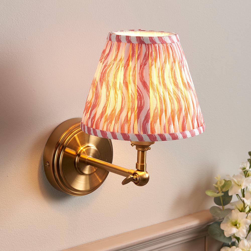 Corven Wall Light in brass with an adjustable knuckle arm and pink Ripple-pattern fabric shade, mounted on a light wall beside a flowering plant.
