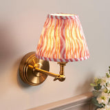 Corven Wall Light in brass with an adjustable knuckle arm and pink Ripple-pattern fabric shade, mounted on a light wall beside a flowering plant.