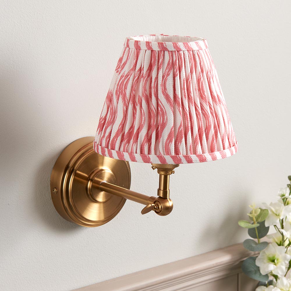 Corven Wall Light in brushed brass with an adjustable knuckle arm and pleated pink and white ripple-pattern fabric shade, shown mounted on a light grey wall.
