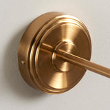 Corven Wall Light in brushed brass with stepped circular backplate and linear arm, close-up on a plain light grey wall.