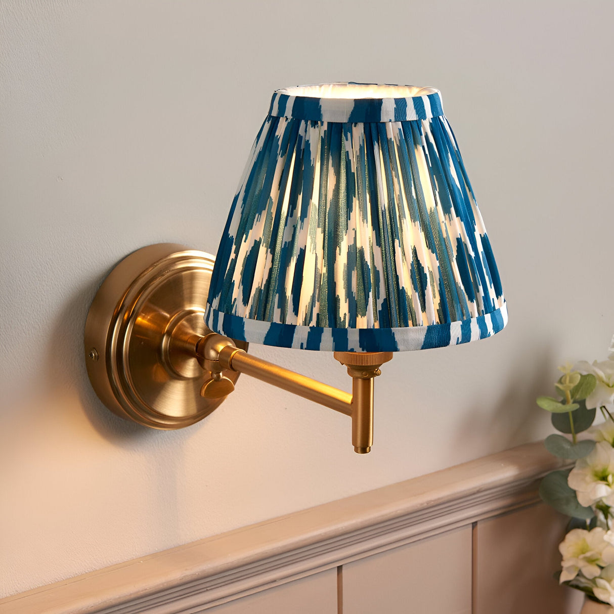 Cliffwood Ikat wall light with brushed brass adjustable arm and swing-arm bracket, pleated blue and white Ikat-print fabric shade, mounted on a light grey wall.