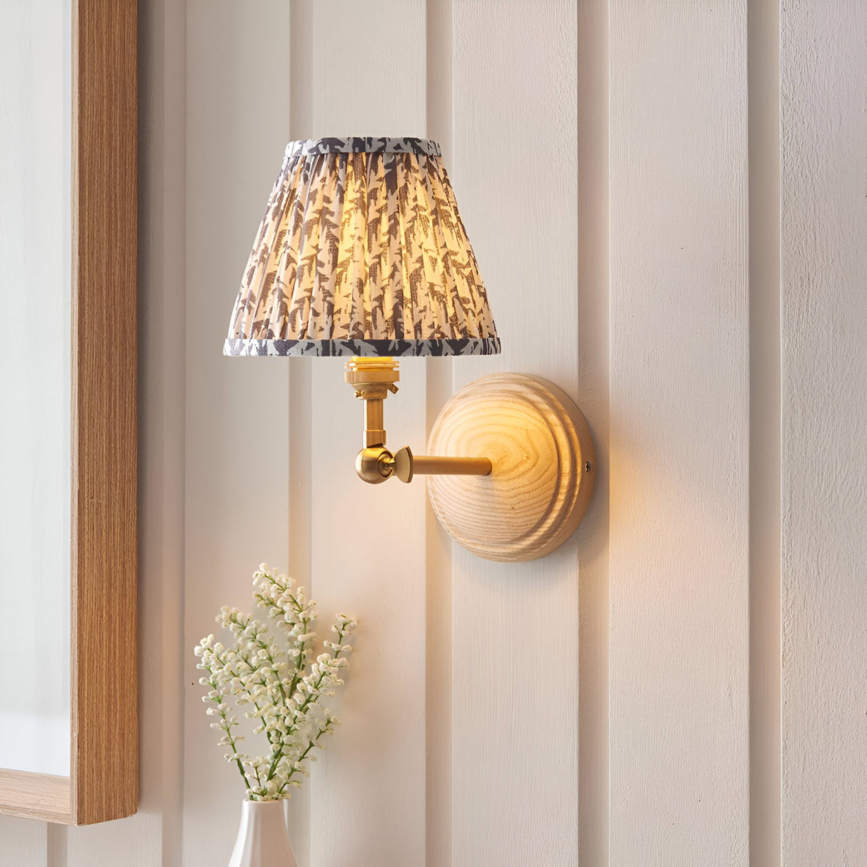 Endon Danemere wall light with adjustable brushed brass arm, natural Ash wood backplate and pleated grey leaf-print shade on a panelled wall.