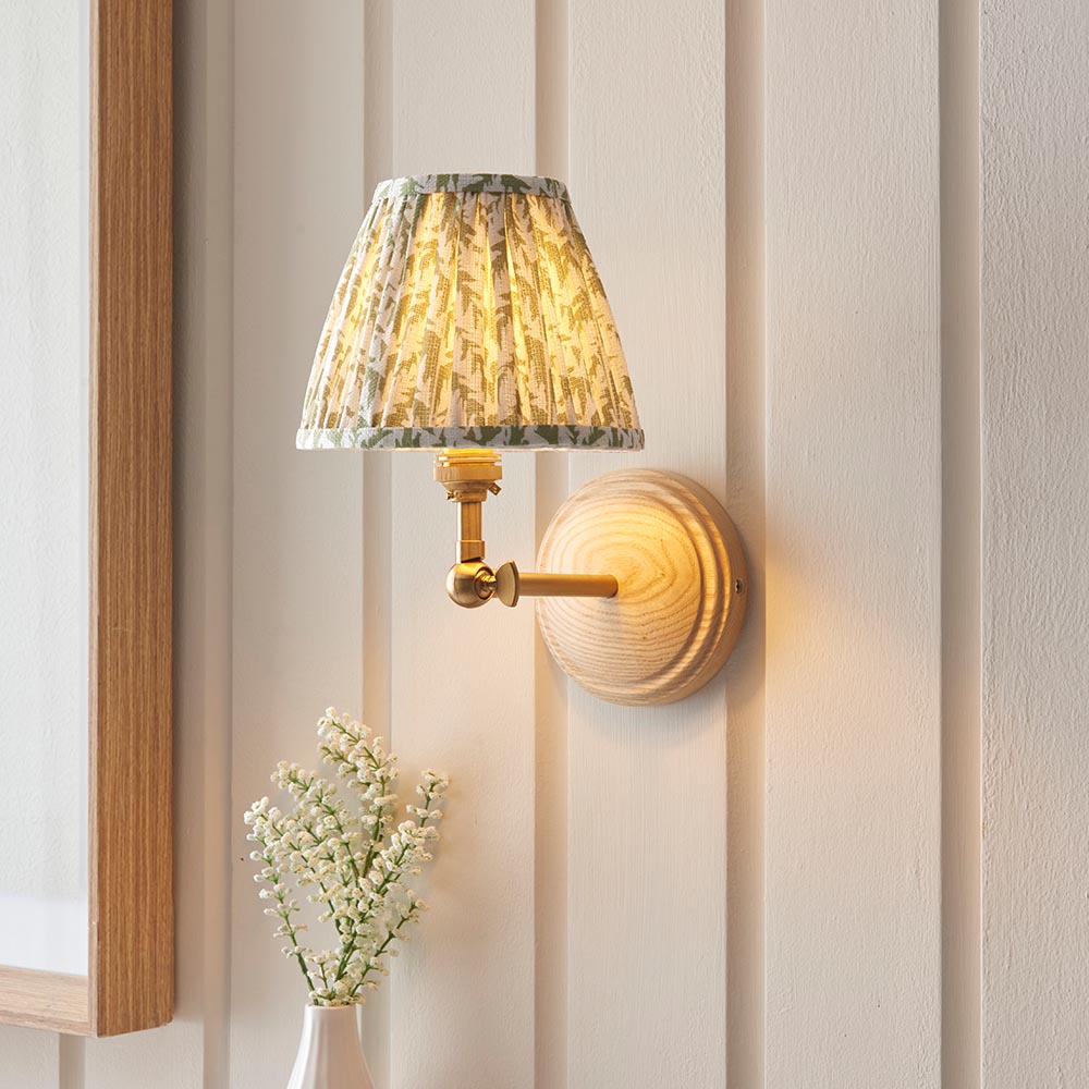 Endon Danemere wall light with natural Ash wood back, brushed brass adjustable arm and green leaf-print faux linen shade, on panelled wall.