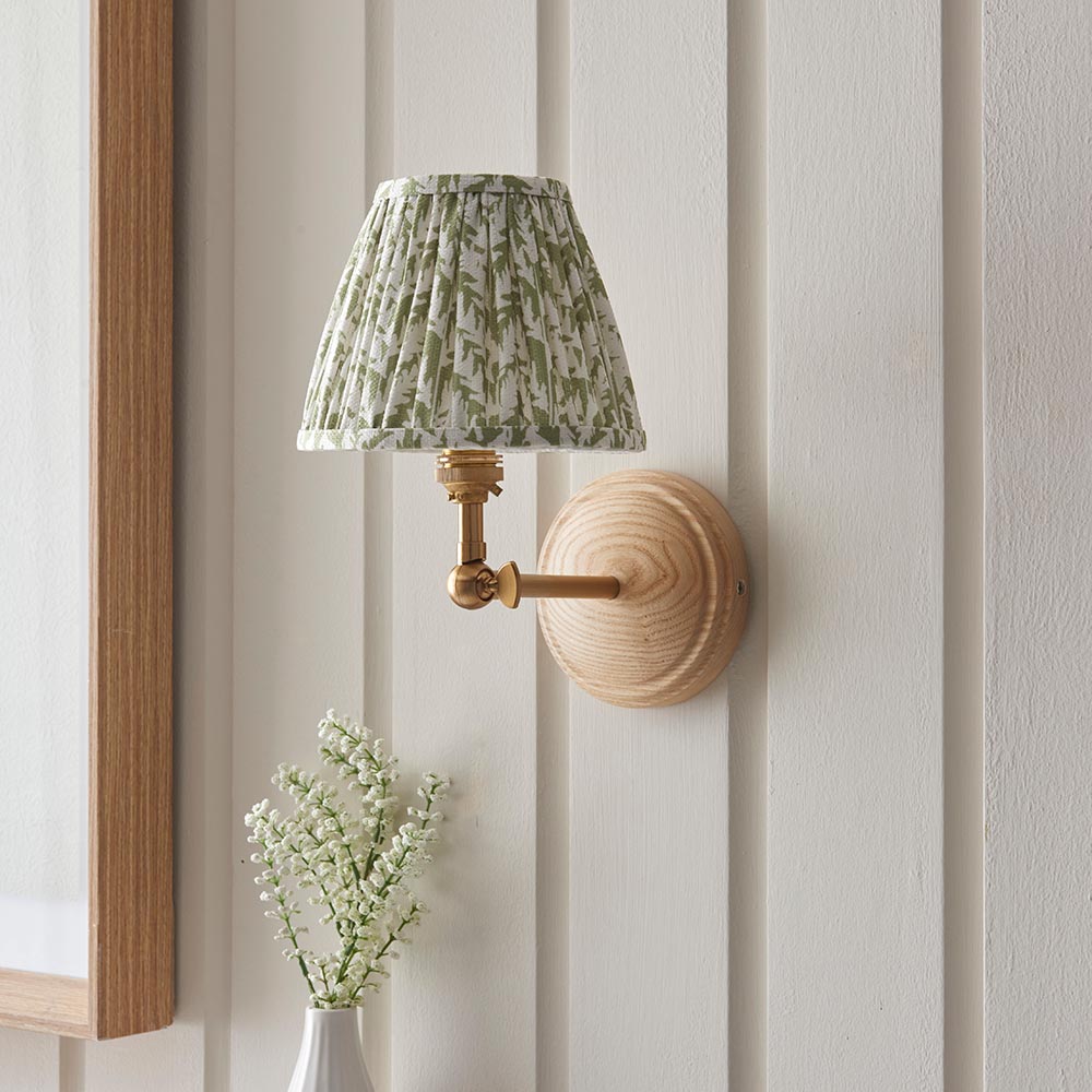 Endon Danemere wall light with natural Ash wood back, brushed aged brass adjustable arm and green leaf-print pleated shade, on panelled wall.