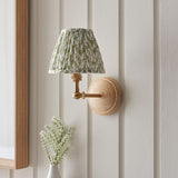 Endon Danemere wall light with natural Ash wood back, brushed aged brass adjustable arm and green leaf-print pleated shade, on panelled wall.