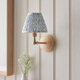 Danemere Wall Light in brass and natural Ash wood with a pleated blue leaf-print fabric shade, shown mounted on a white panelled wall.