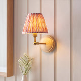 Danemere Ripple wall light with wood backplate, brass arm and adjustable knuckle, tapered pink and orange striped fabric shade, shown lit on a panelled wall.