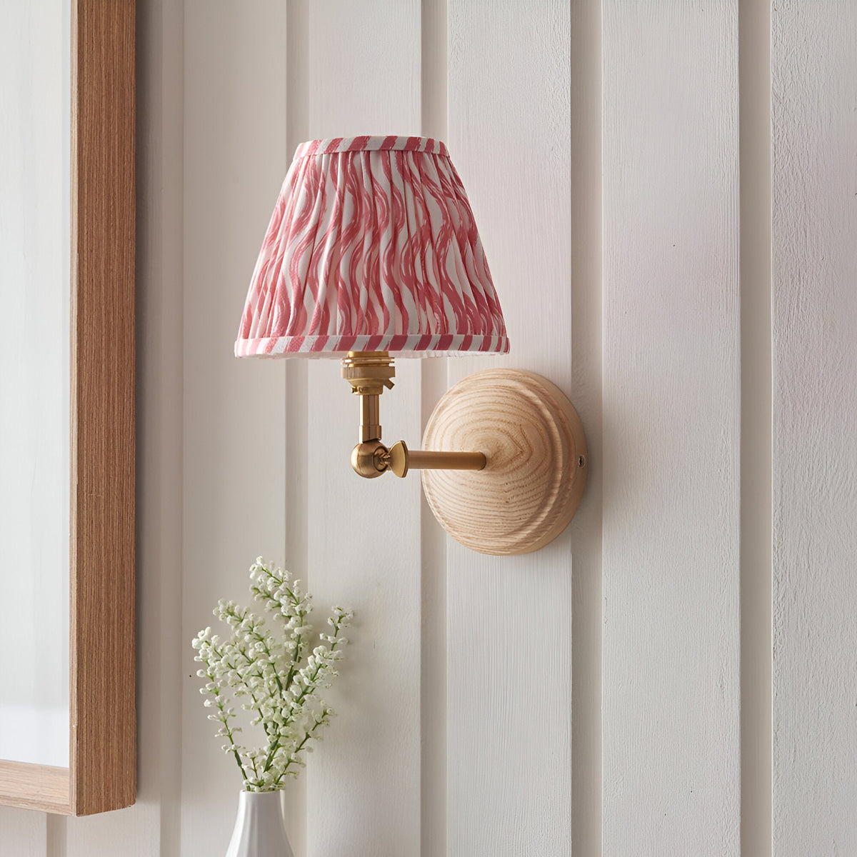 Danemere Ripple wall light with pleated coral pink patterned shade, brass fittings and adjustable knuckle on a natural wood backplate, shown on a panelled wall.