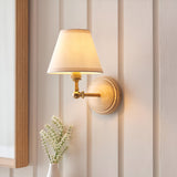 Danemere Ivy wall light with tapered beige fabric shade, brass arm and adjustable knuckle on a round ash wood back plate, mounted on a panelled wall.