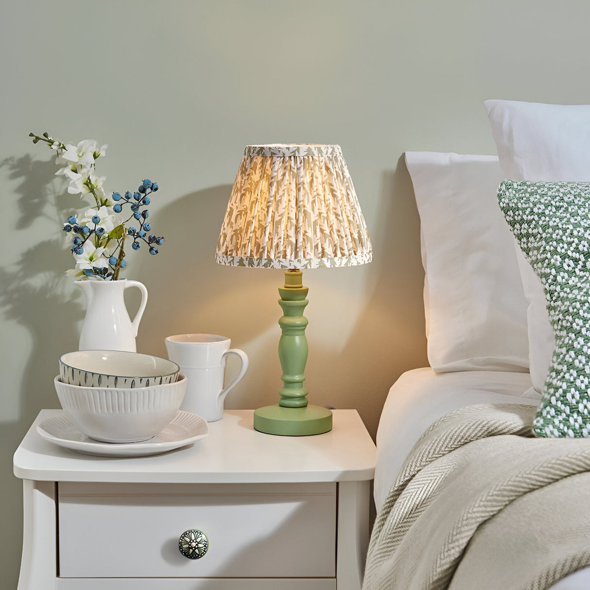 Endon Aldworth Leaf table lamp with green turned wooden spindle base and pleated leaf-print shade on a white bedside drawer beside a bed.