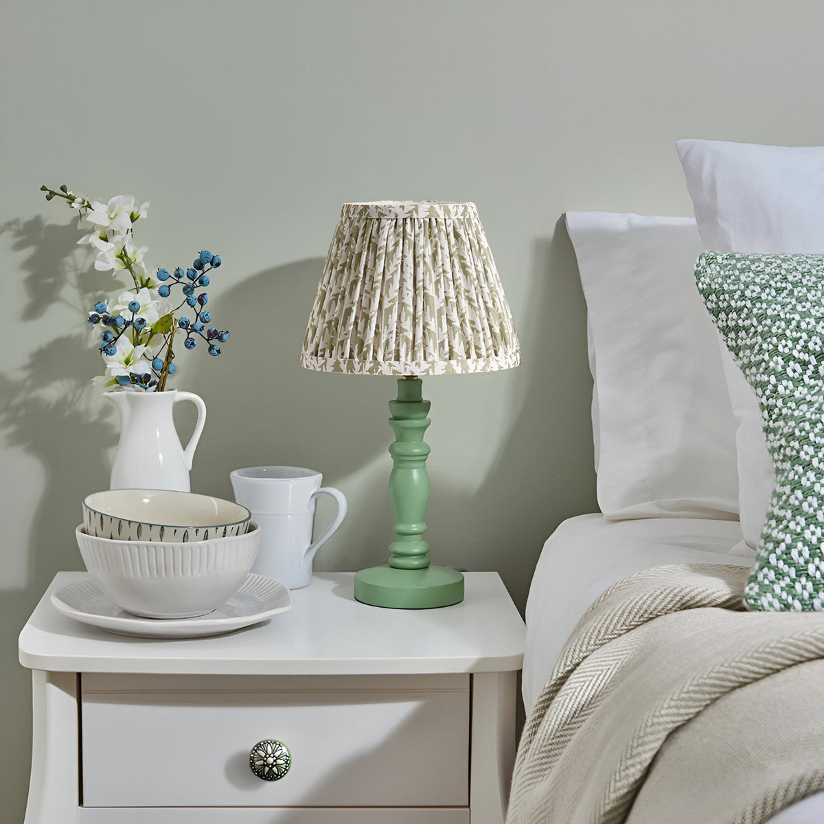 Endon Aldworth Leaf table lamp with green turned spindle base and pleated leaf-print shade on a white bedside table styled with jug, bowls and mug.