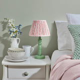Endon Aldworth Leaf table lamp with green turned wooden spindle base and pink leaf-print pleated shade, styled on a white bedside table beside a bed.