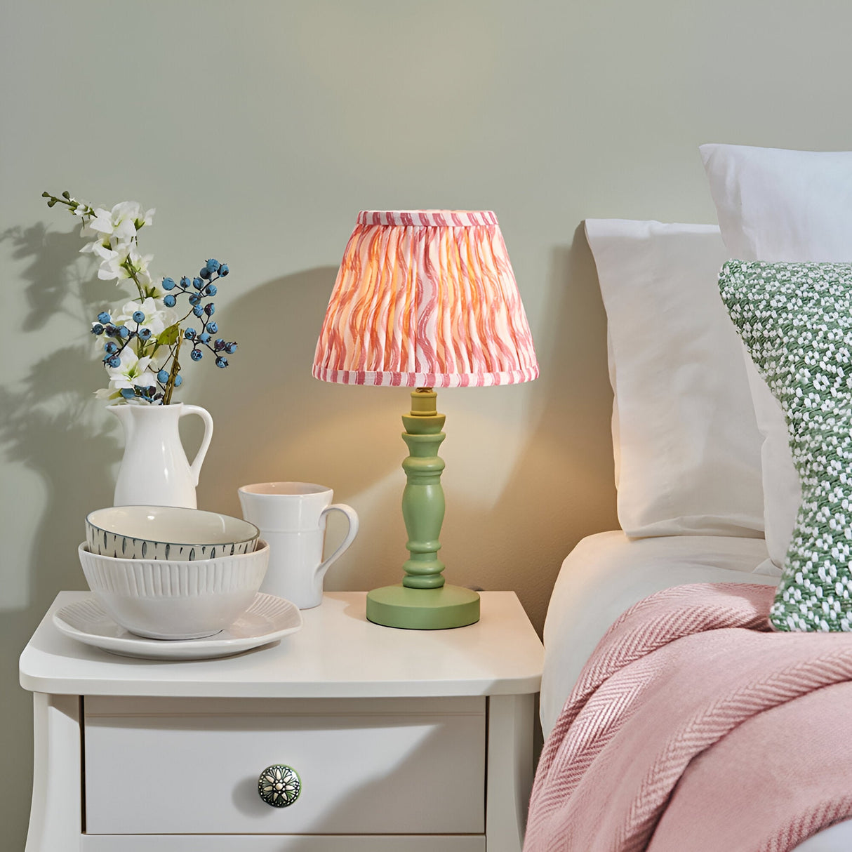 Endon Aldworth Ripple table lamp with green turned wood spindle and coral pink pleated ripple shade on a white bedside drawer beside a bed.