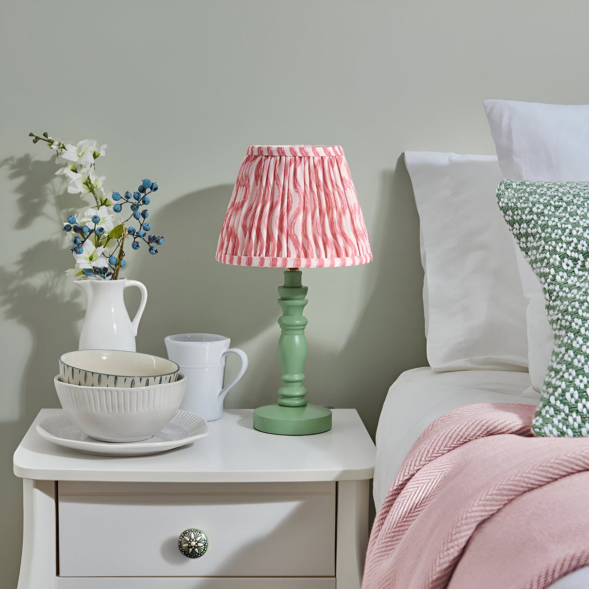 Endon Aldworth Ripple table lamp with green turned spindle base and coral pink pleated ripple-print shade, on a white bedside drawer in a bedroom.
