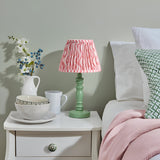 Endon Aldworth Ripple table lamp with green turned spindle base and coral pink pleated ripple-print shade, on a white bedside drawer in a bedroom.