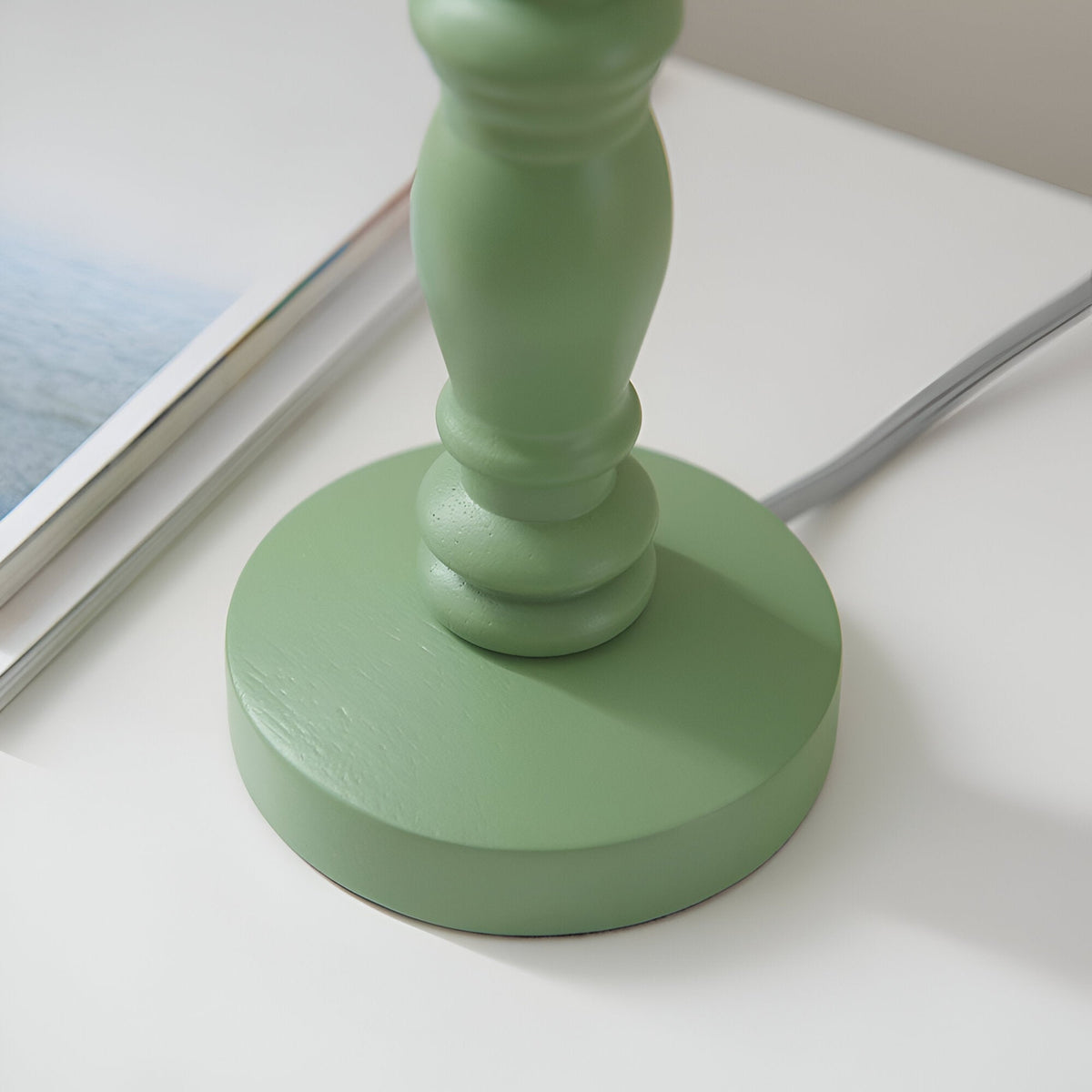 Close-up of the green satin painted turned wooden base of the Endon Aldworth Ripple table lamp with grey braided flex, beside a magazine.