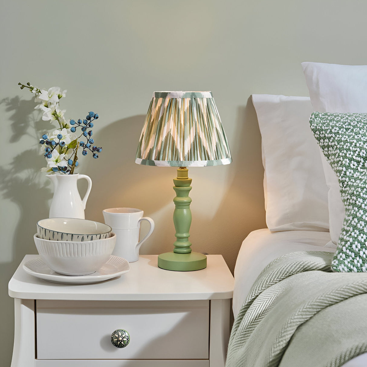 Endon Aldworth table lamp with green turned wood spindle base and pleated green, grey and cream zigzag shade on a white bedside drawer beside a bed.
