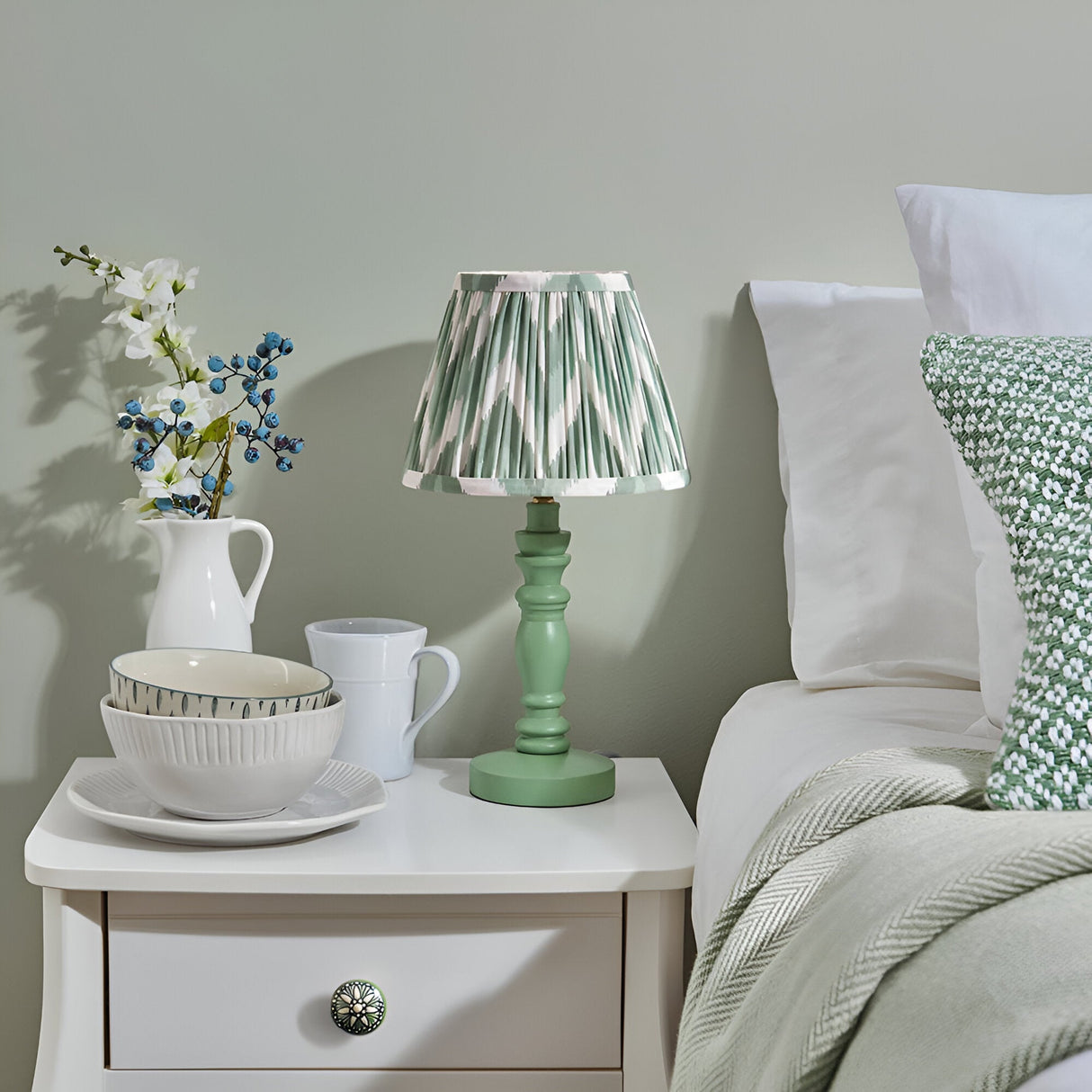 Endon Aldworth table lamp with green turned wooden spindle base and pleated green zigzag shade on a white bedside table beside a bed.