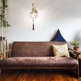 A cozy living room includes a brown leather sofa embellished with a patterned cushion and a blue throw pillow. The Daley Wall Light in brass, with its antique finish, hangs above, providing a warm glow. A potted plant sits on the wooden floor next to the sofa.