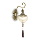 The Daley Wall Light - Brass features a stunning wall-mounted design with a clear glass shade, intricate metal detailing, and a decorative tassel at its base. It is finished in an antique style with elegant curves and vintage accents, casting a soft, warm glow.