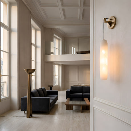 A modern, minimalist living room with high ceilings, large windows, black sofas, a wooden coffee table, a tall brass floor lamp, and the Elystria LED Vertical Stone Alabaster Wall Light in soft white and brass accents, all in neutral tones.