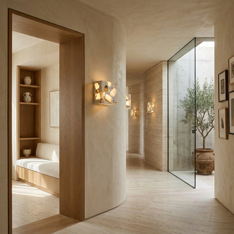 A modern hallway with curved beige walls, wood accents, built-in seating, framed art, and an olive tree by a floor-to-ceiling window is softly lit by the Terrano 1 Light Cube Travertine Wall Light in light beige.