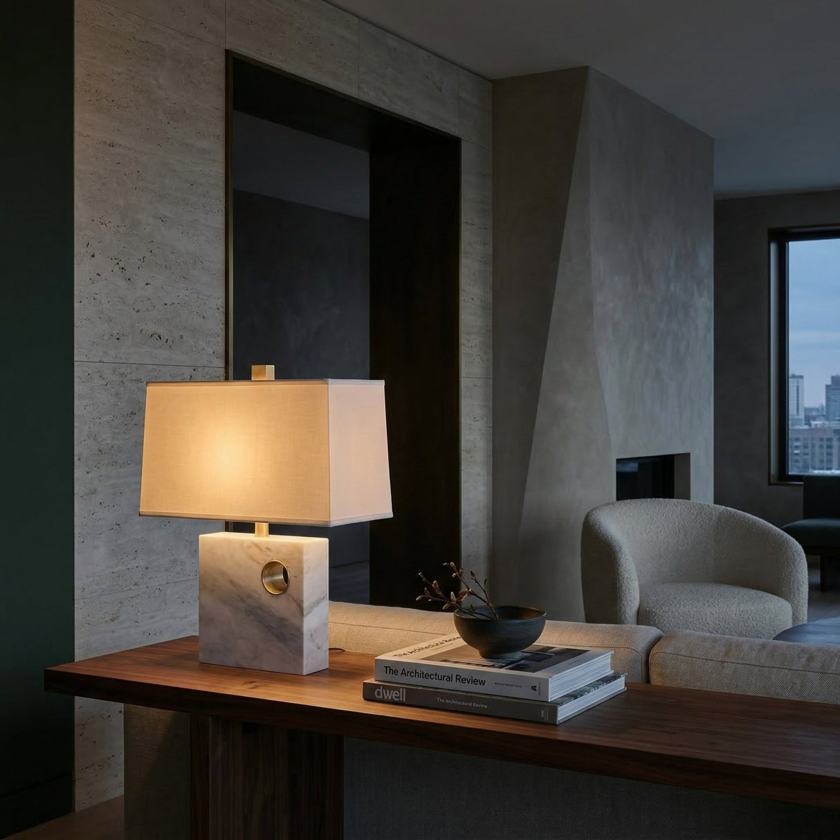 A modern living room with a wooden console table topped with the Corvella 1 Light Pierced Marble Table Lamp in brass and white, stacked books, a small black bowl, and a light armchair by a window overlooking the city.
