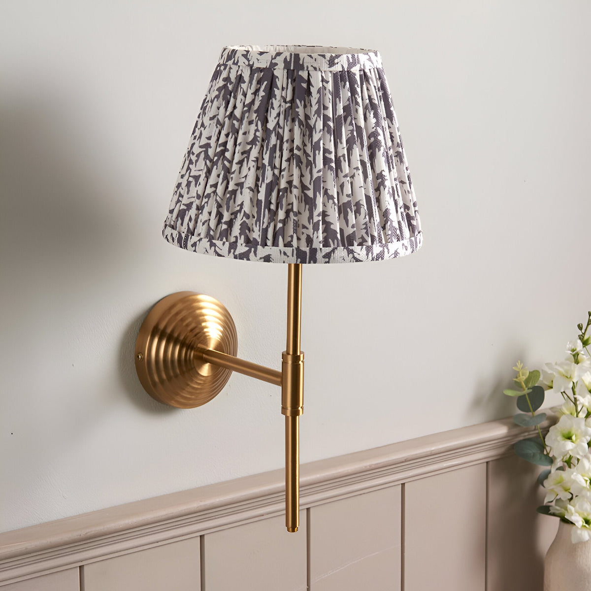 Castleigh Leaf wall light in grey with brushed brass stepped back plate and T-shaped arm, pleated tapered fabric shade, mounted on a light grey wall.